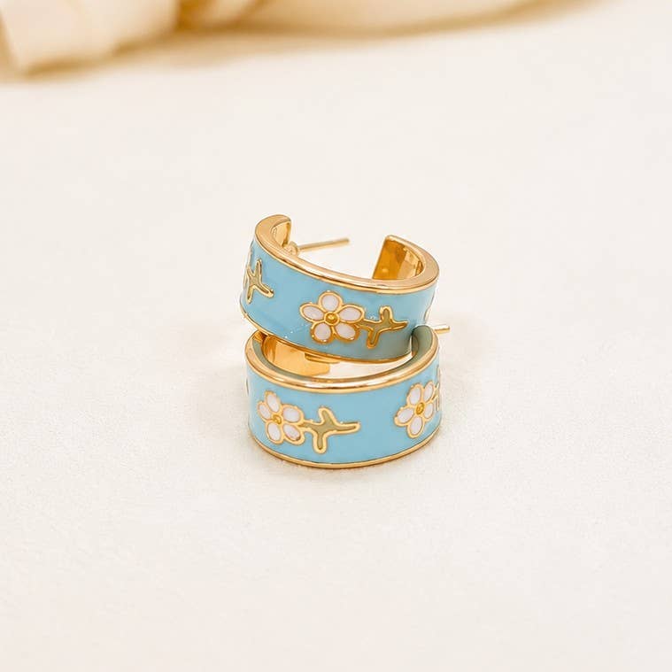 A pair of gold hoop earrings with light blue enamel and white cherry blossom motifs, displayed on a soft cream-colored surface.