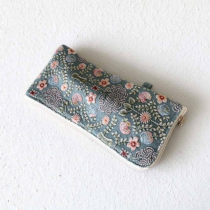 A rectangular clutch purse with an intricate floral and paisley pattern in muted blue, pink, and green tones, featuring a white border and a small gold clasp, resting on a textured white wall.