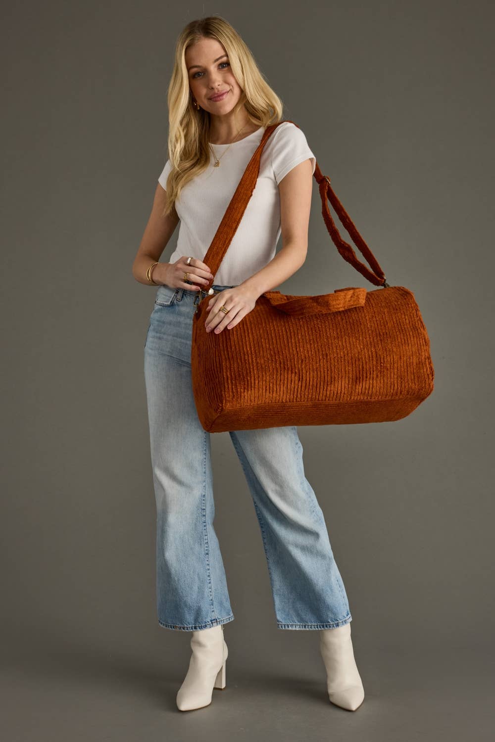 A person holding a large rust-orange corduroy duffel bag with top handles and a long shoulder strap against a plain gray studio background.