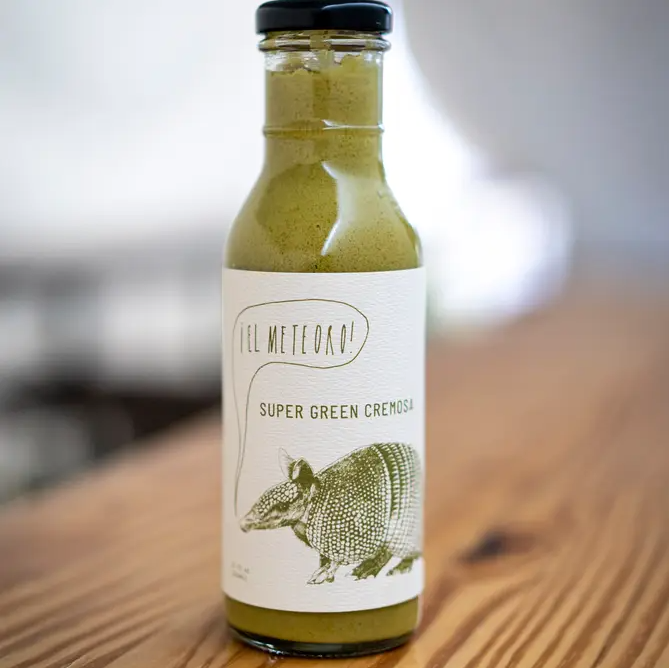 A bottle of Ill Meteoro Super Green Crema on a wooden table with a blurred indoor background. The bottle is filled with a green sauce and has a white label with a green armadillo illustration.
