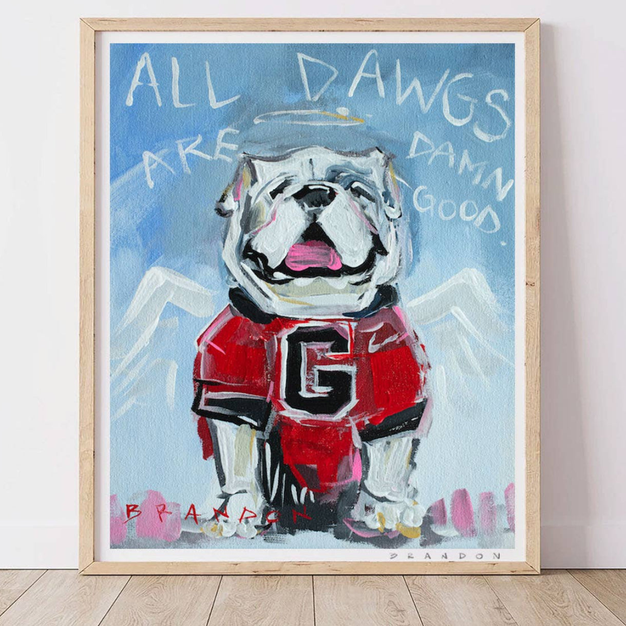 A framed art print featuring a painted bulldog wearing a red football jersey with the letter G, set against a blue sky with white text reading 'ALL DOGS ARE DAMN GOOD.' The print is displayed in a light wood frame leaning against a white wall on a light wood floor.