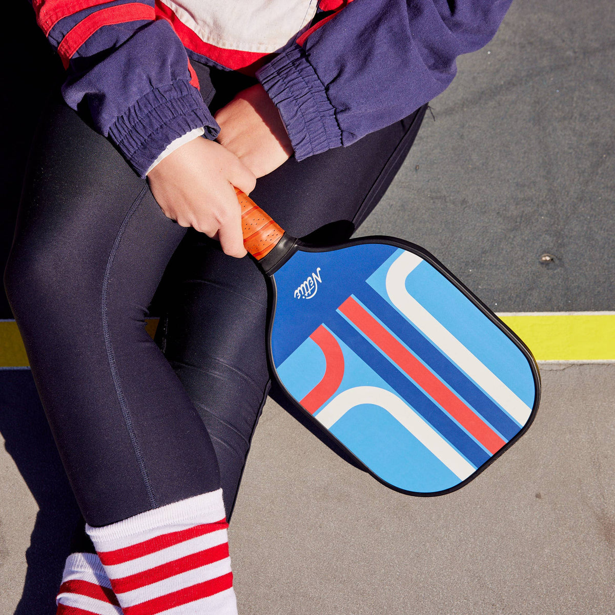 A person sitting outdoors on a court holding a pickleball paddle with a blue, red, and white geometric pattern and an orange handle.