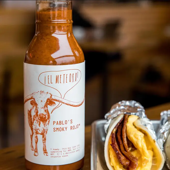 A bottle of Pablo's Smoky Rojo hot sauce from El Meteoro, featuring a white label with a brown steer illustration, shown next to a taco filled with eggs and bacon on a tray.