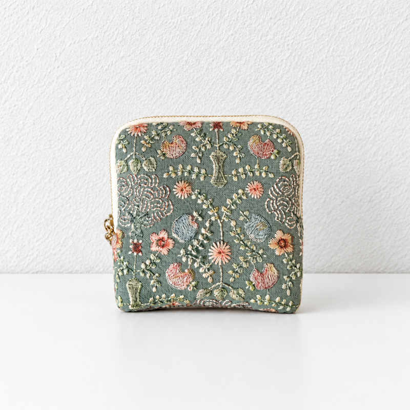 A small zippered pouch with an intricate floral and botanical embroidery pattern on a muted sage green fabric, featuring a cream zipper and gold-tone hardware, set against a white textured wall and white surface.