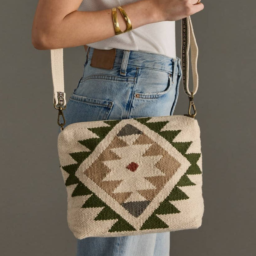 A woman wearing a white t-shirt and blue jeans carries a woven crossbody bag with a Southwestern geometric pattern in beige, brown, and green.