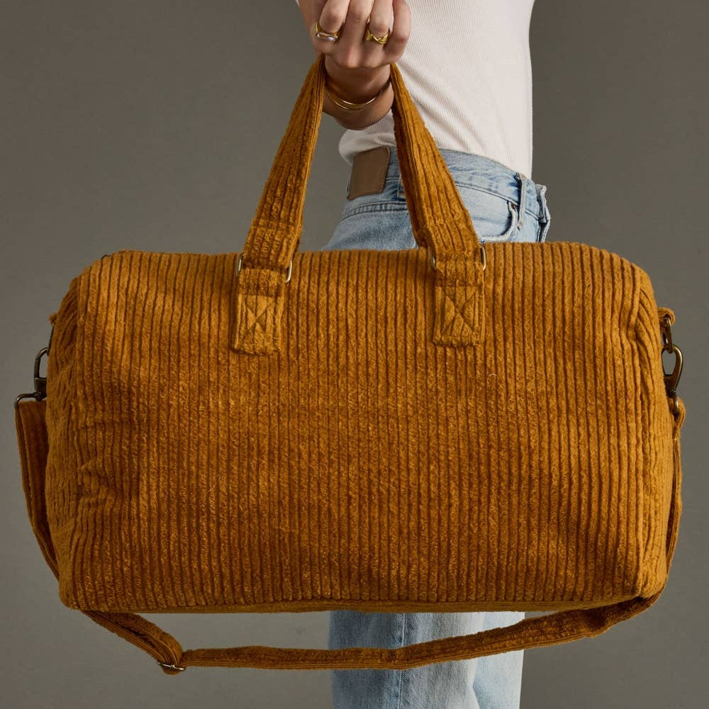Person holding a mustard yellow corduroy duffel bag against a gray background