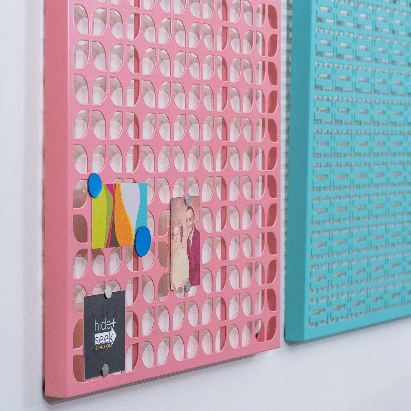 Pink and turquoise plastic grid wall organizers mounted on a white wall, with small colorful items pinned to the pink one.