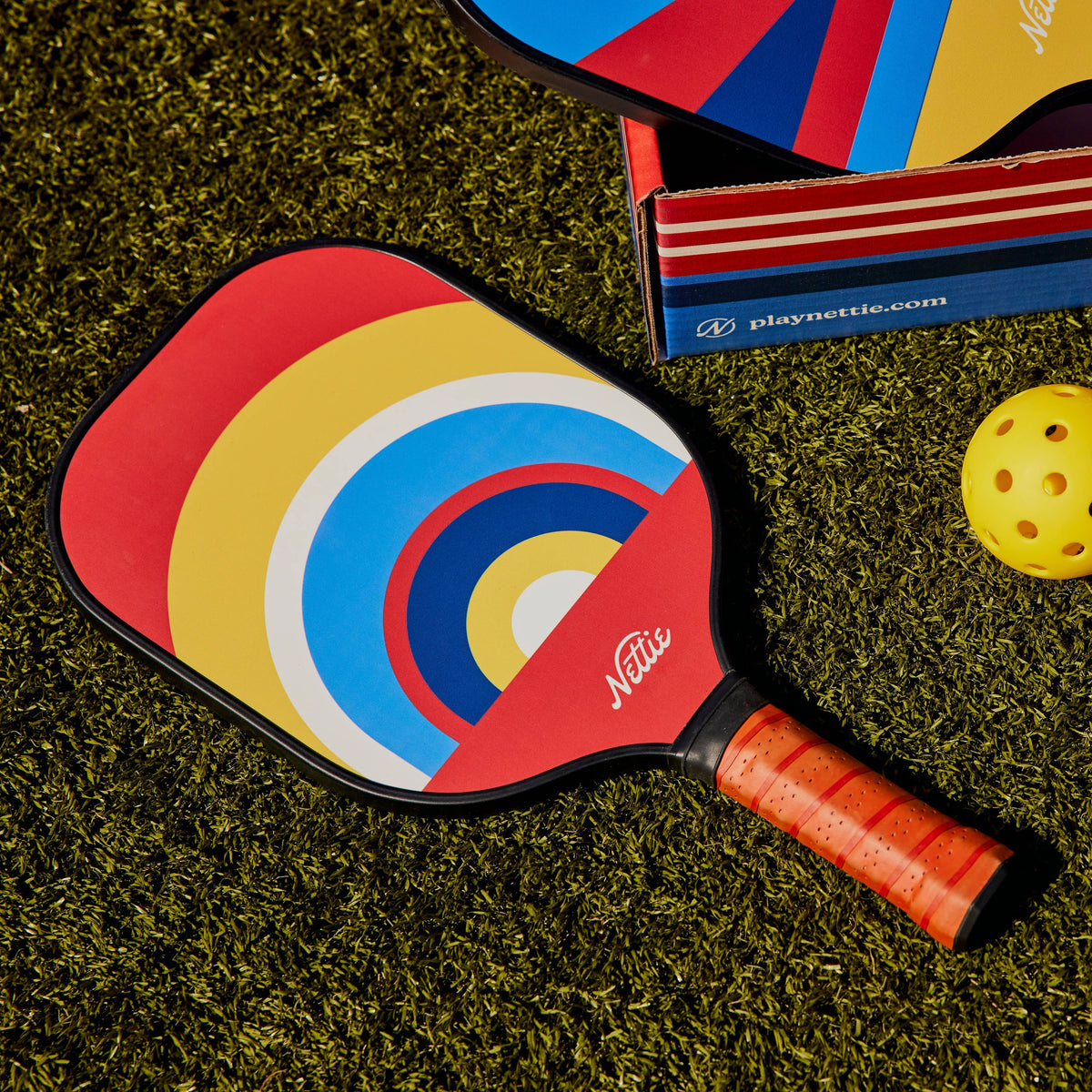 A colorful pickleball paddle with a rainbow target design, a wrapped orange handle, two yellow pickleballs, and part of the product box on a green turf-like surface.
