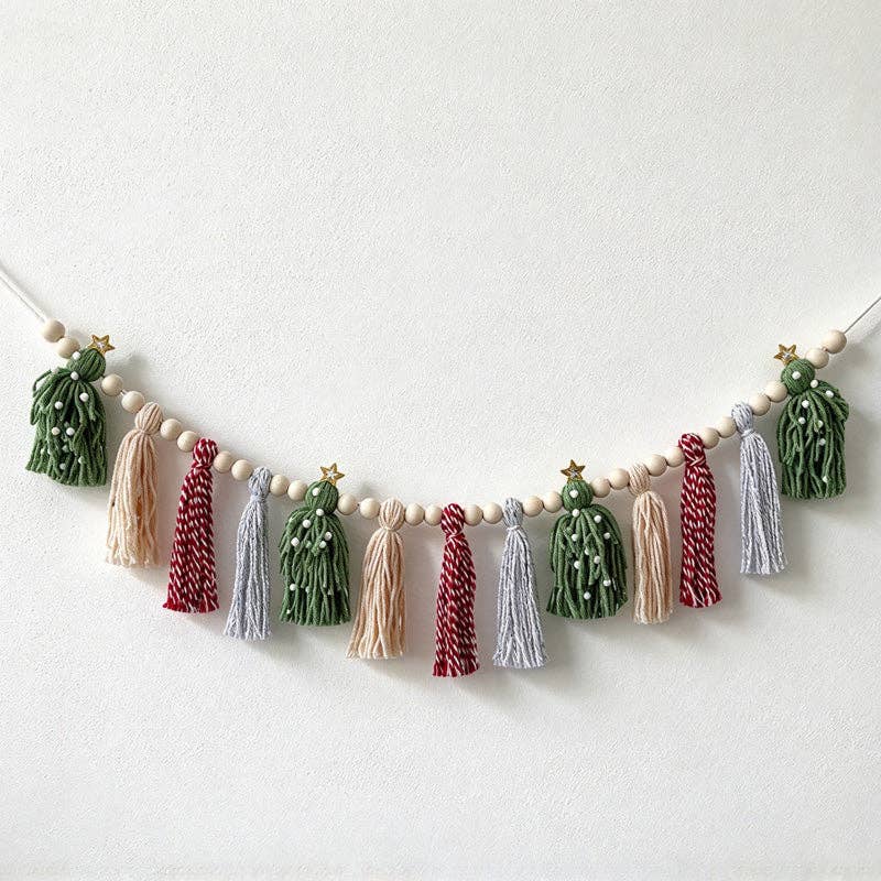 A festive Christmas garland featuring miniature green Christmas trees with gold star toppers, hanging tassels in green, beige, burgundy, and light gray, and small white wooden beads, strung on a white cord against a plain off-white wall.