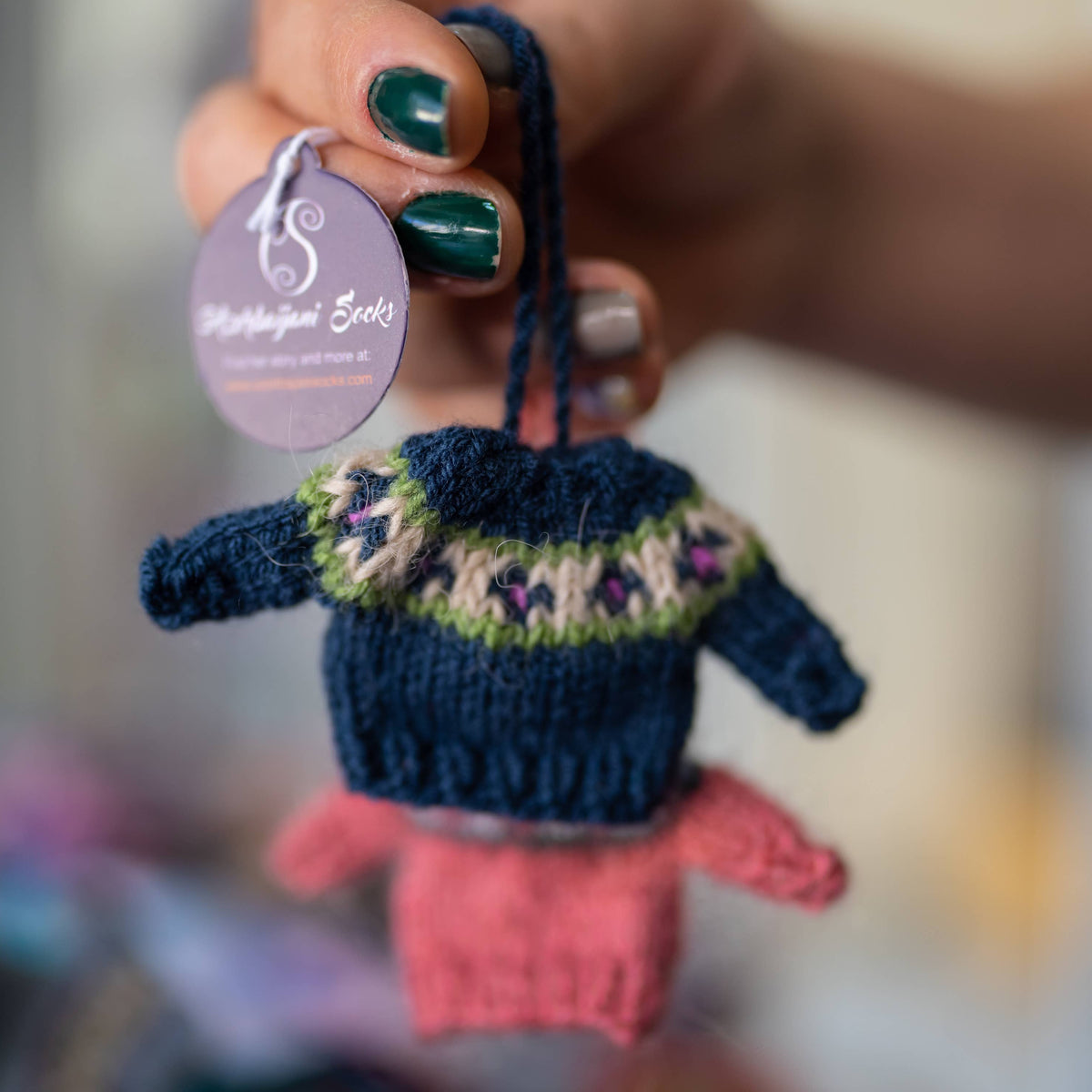 A hand holds a tiny knitted winter sweater, featuring a dark blue, green and purple patterned print.