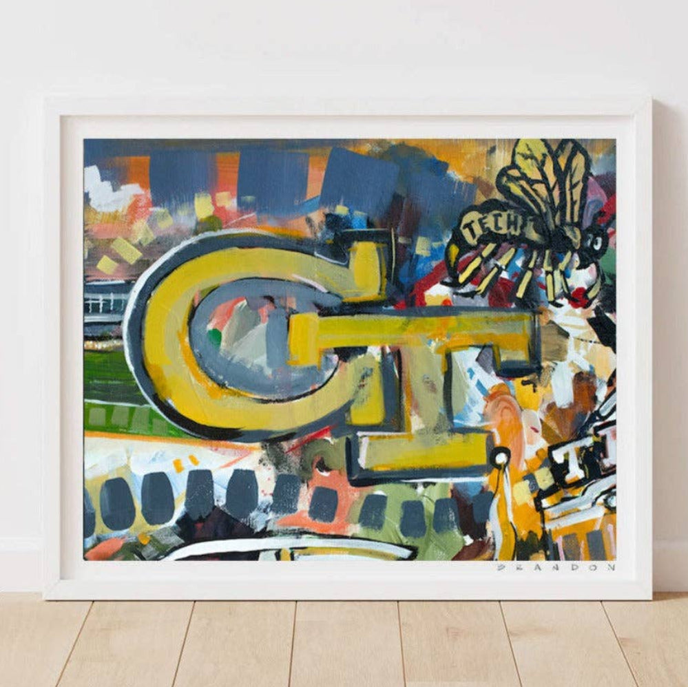 A framed abstract art print leaning against a white wall on a light wood floor. The artwork features a large yellow and gray stylized letterform over a multicolored abstract background with blue, green, orange, black, and white elements.