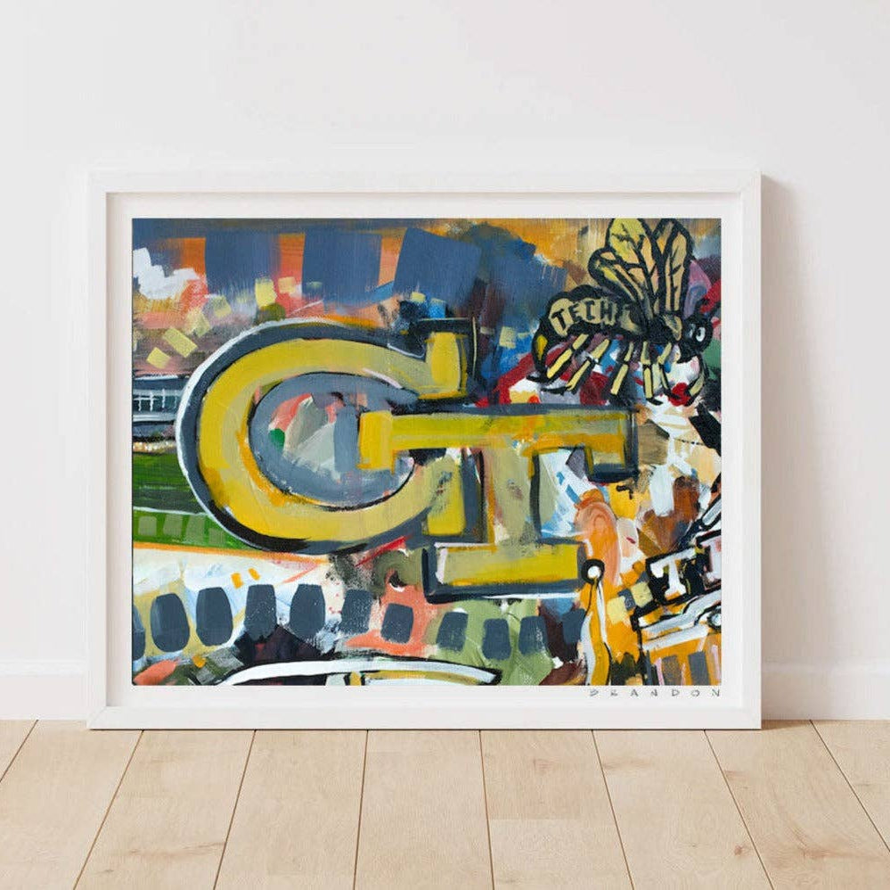 A framed abstract art print leaning against a white wall on a light wood floor. The artwork features a large yellow and gray stylized letterform over a multicolored abstract background with blue, green, orange, black, and white elements.