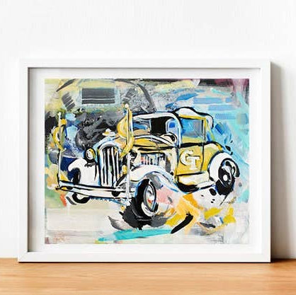 A framed abstract art print depicting a vintage yellow and white truck, leaning against a white wall on a light wood surface.