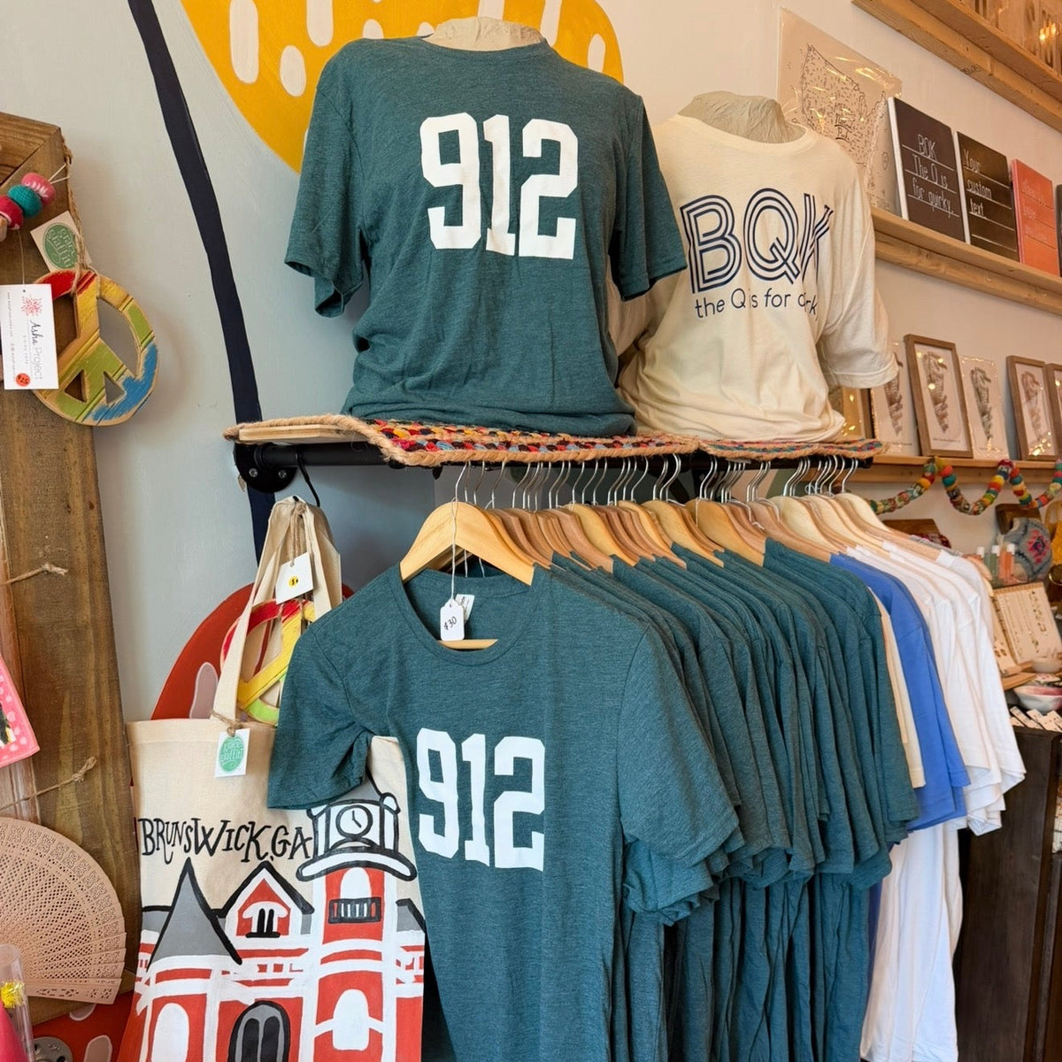 A retail display of t-shirts, with a dark teal heathered shirt featuring the white printed number '912' hanging prominently in the foreground, alongside other shirts on wooden hangers and mannequins.