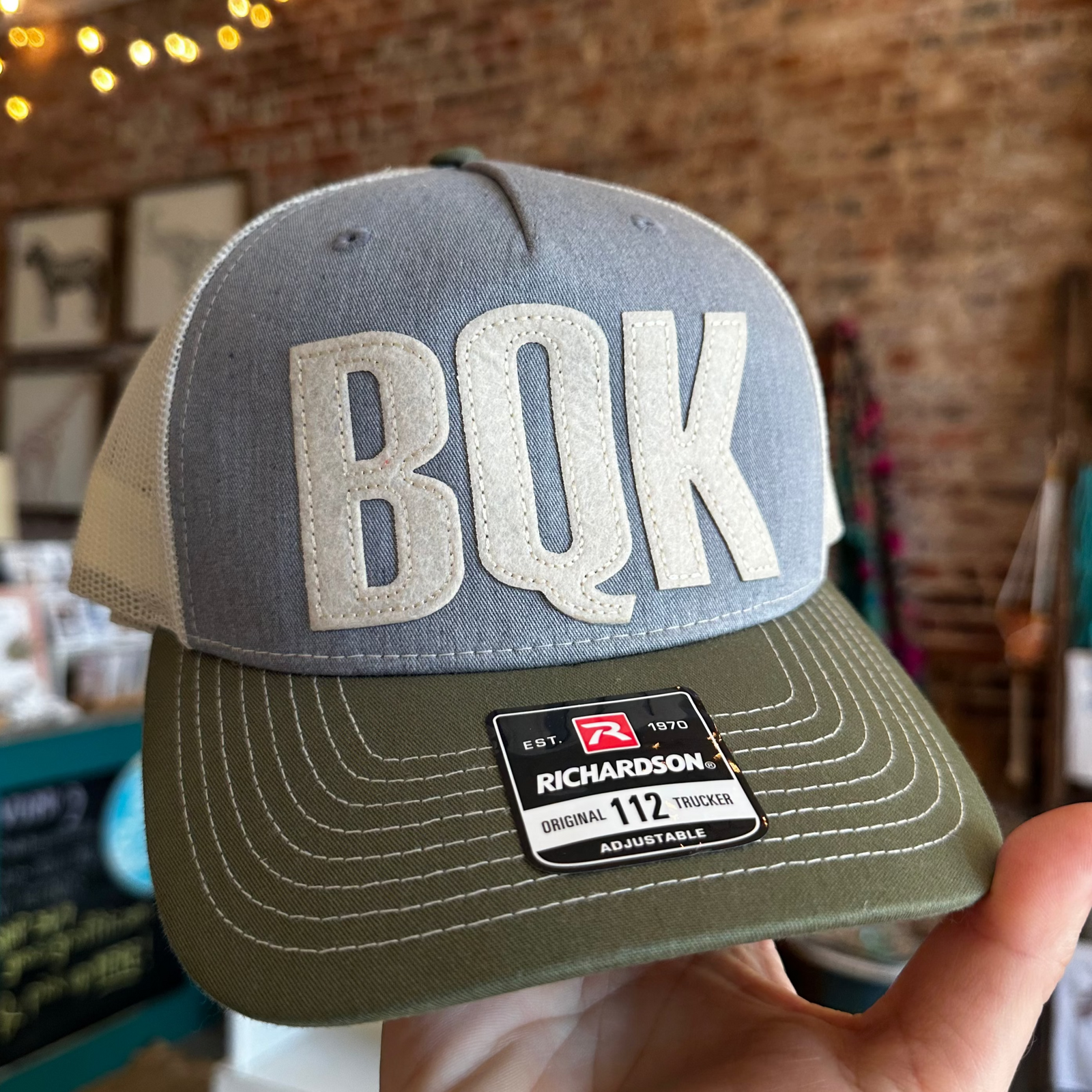 A hand holding a five-panel twill trucker cap with 'BQK' text on it, predominantly grey with green visor and white stitching details.