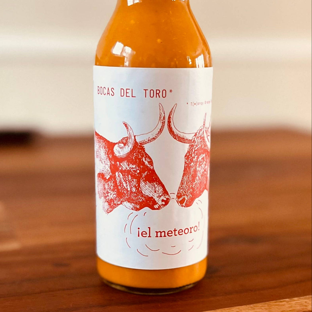A small glass bottle of orange-red sauce with a black cap and a white label featuring red bull illustrations, standing on a wooden surface against a softly blurred indoor background.