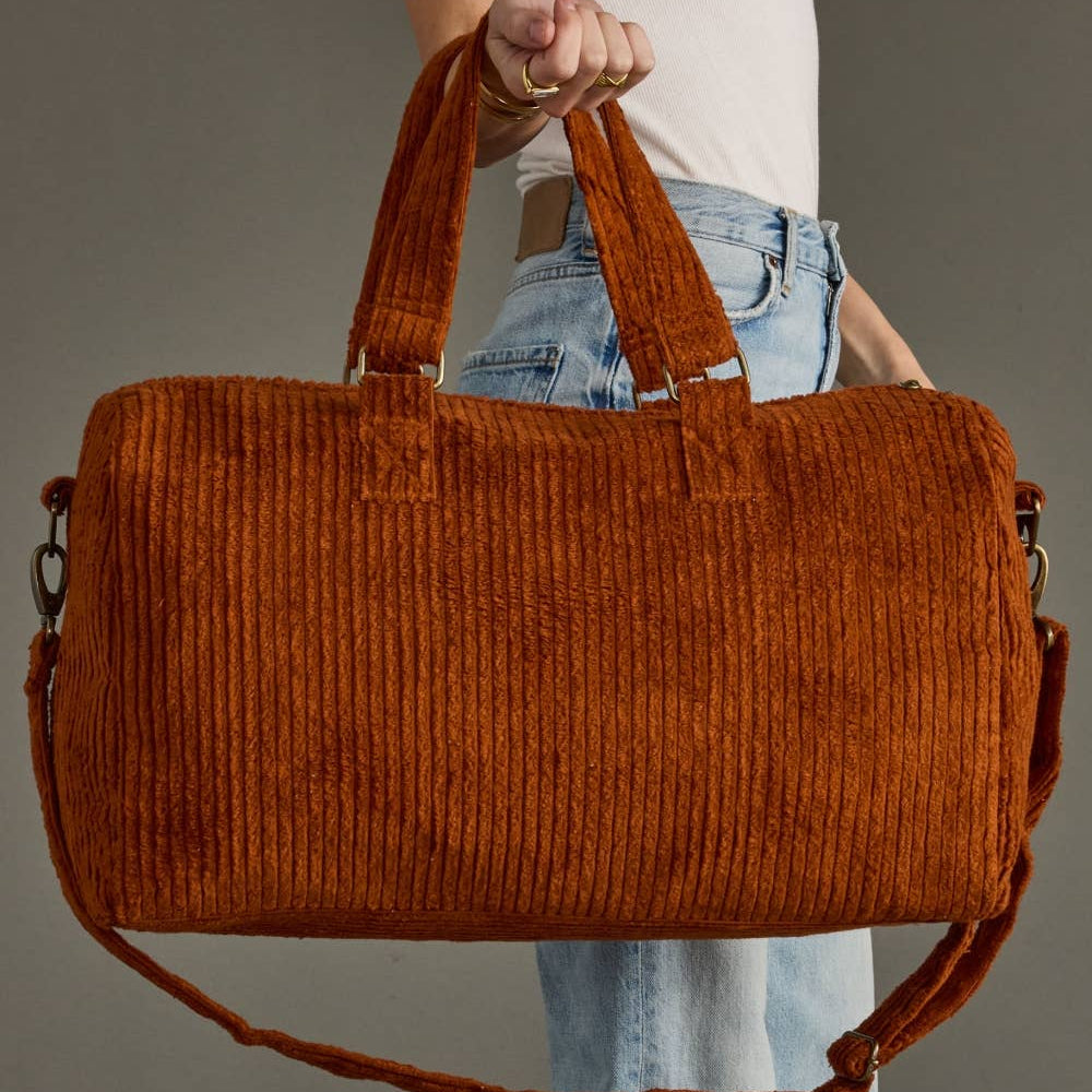 A person holding a large rust-orange corduroy duffel bag with top handles and a long shoulder strap against a plain gray studio background.