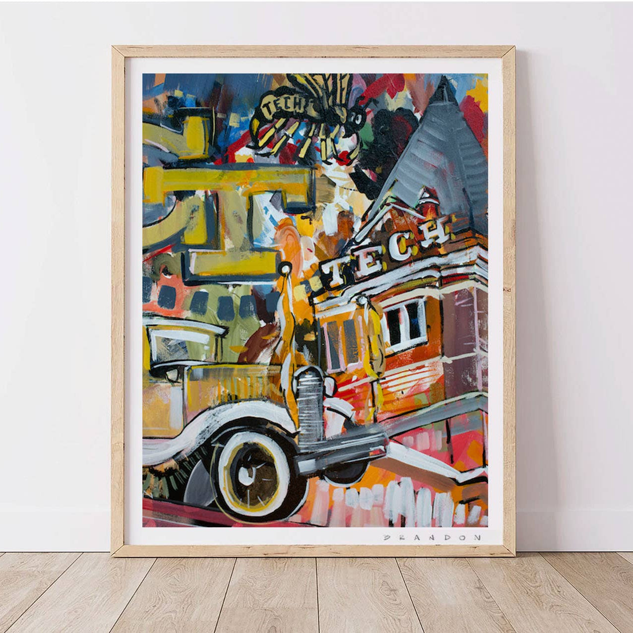 A framed abstract art print leaning against a white wall on a light wood floor. The artwork features a colorful, painterly depiction of a vintage car and building with graffiti-style lettering.