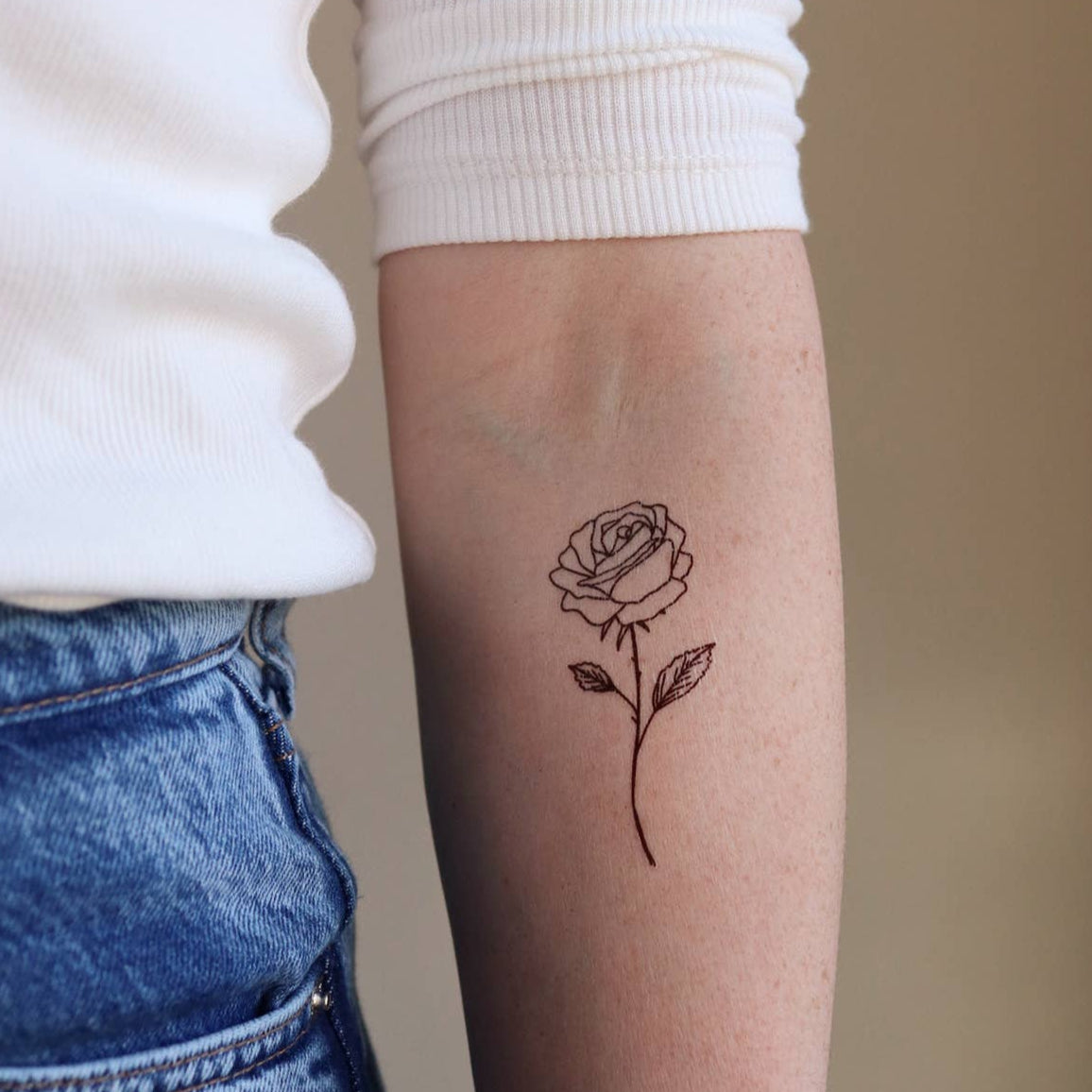 Person wearing a white long-sleeve shirt and blue jeans with a small rose tattoo on their arm.