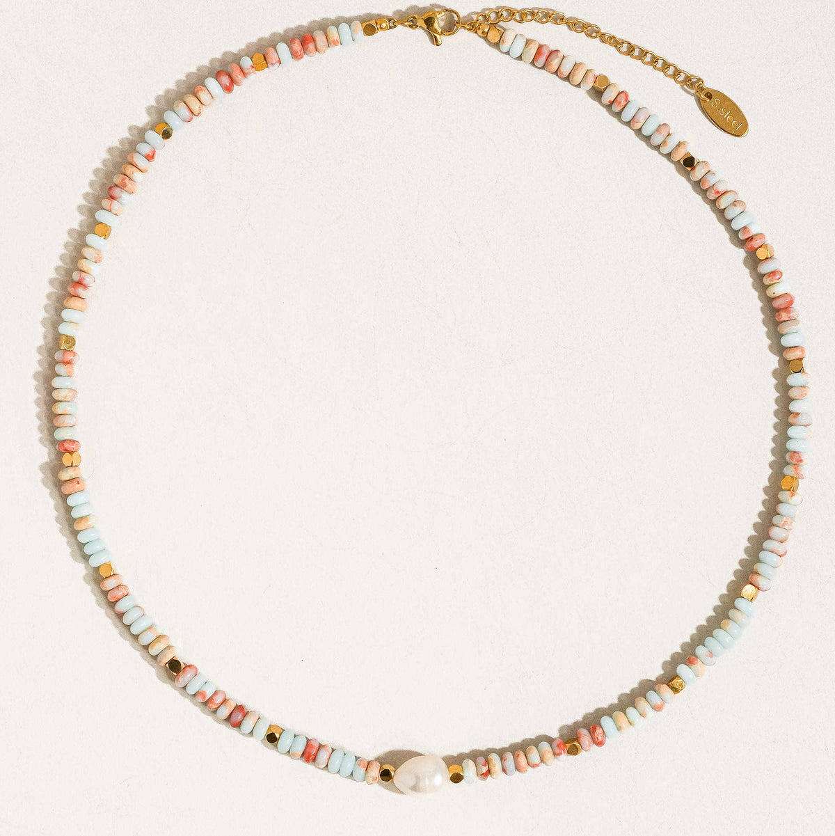A delicate beaded necklace with small pastel-colored cylindrical beads in pink, orange, yellow, and blue, accented by a white cowrie shell and gold-tone hardware, displayed on a textured off-white background.