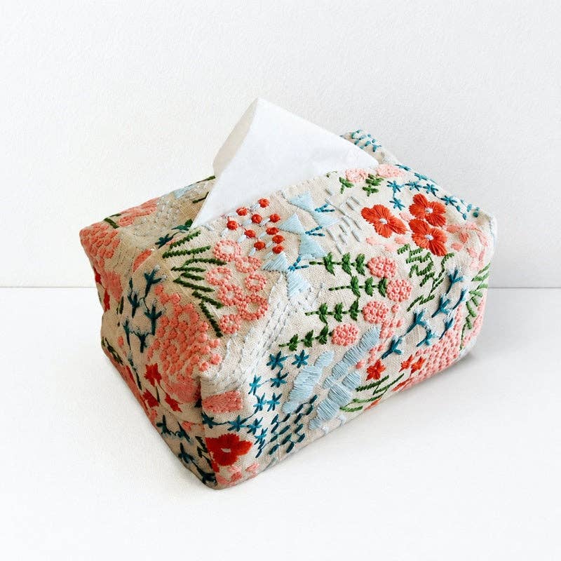A square tissue box cover with a colorful floral and leaf embroidery pattern on a cream background, with a white tissue paper partially pulled out at the top, set against a plain white background.