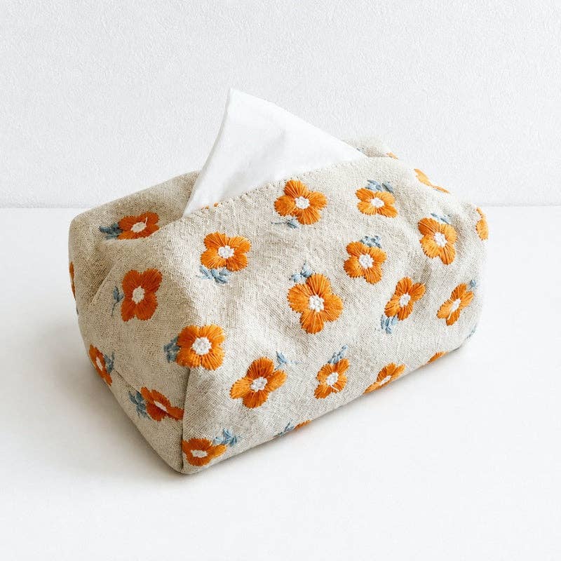 A fabric tissue box cover with an embroidered orange and white floral pattern on a light beige background, with a white tissue peeking out from the top, set against a plain white background.