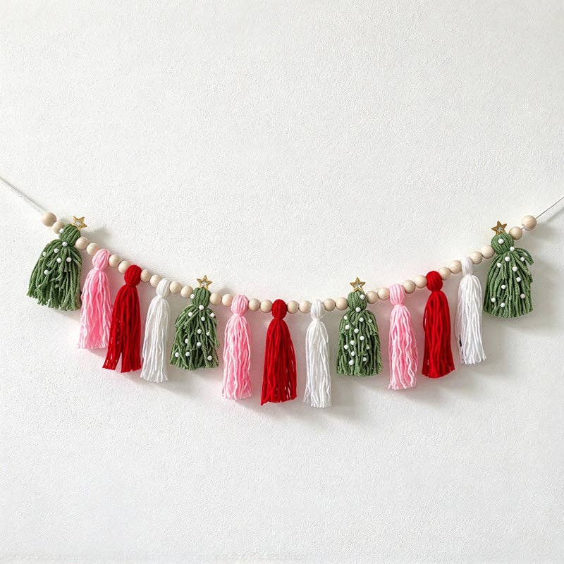 A festive Christmas garland featuring tinsel tree ornaments in green and white, accented with red and pink tassels, white wooden beads, and small gold star accents, hanging against a plain white wall.