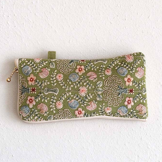 A rectangular fabric pouch with a green base and an intricate multicolor floral and paisley pattern, featuring a gold-tone zipper pull and a small green tab at the top, shown against a white textured wall.