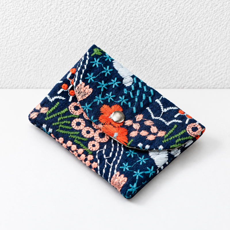 A small fabric pouch or clutch with a dark navy base and colorful embroidered floral and geometric patterns, featuring a flap closure with a silver metal snap button, photographed on a white surface against a white textured wall.