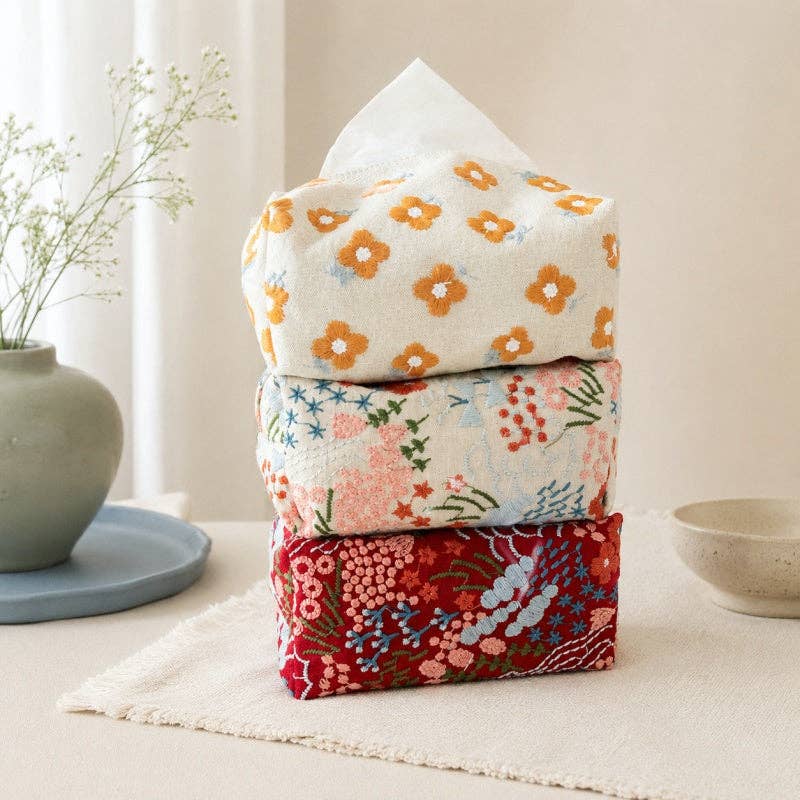 Three stacked fabric tissue boxes with floral patterns in orange, pink, and red, set on a light beige table runner with a vase of flowers and a bowl in the background.